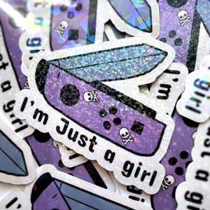 May include: A collection of holographic stickers featuring a purple game controller design with the text "I'm Just a girl". The stickers have a glittery effect and include skull and crossbones graphics.