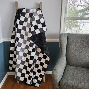 Op de afbeelding: Een quilt met een herhalend dambordpatroon in tinten zwart, wit en grijs. De stof toont een raster van vierkanten met zichtbare stiklijnen. Eén kant toont het ruitjesontwerp, terwijl de achterkant volledig zwart van kleur is.