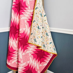 May include: A pink and white quilt with a geometric star pattern. The quilt is folded and draped over a wooden ladder.