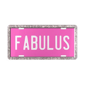 Könnte beinhalten: Rechteckiges Nummernschild mit leuchtend rosa Hintergrund und dem Wort "FABULUS" in fetten weißen Buchstaben. Der Rand ist silberfarben mit Glitzer. Das Schild hat vier Befestigungslöcher.