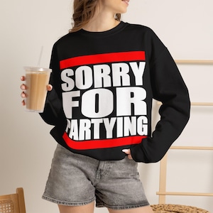 以下が含まれることがあります： 黒の長袖スウェットシャツ。中央には太字のサンセリフ体で「SORRY FOR PARTYING」という白い文字がプリントされ、その上下を赤い水平のストライプが囲んでいます。グレーのデニムショートパンツと組み合わされています。