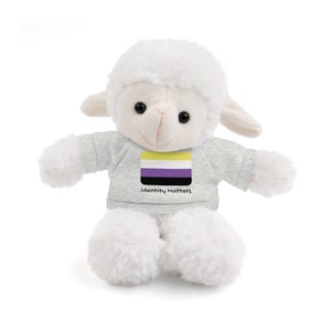 May include: A white plush lamb wearing a gray t-shirt. The t-shirt displays the non-binary pride flag and the words "Identity matters". The lamb has black eyes and a pink nose. The lamb's fur is soft and fluffy.