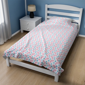 May include: White twin bed with a patterned duvet cover and pillow. The pattern features pink cat faces and blue coffee bean shapes on a white background. The bed frame is white, and the room has blue walls and a wooden floor.