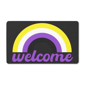 Puede incluir: Una alfombra rectangular con fondo negro presenta un diseño de arcoíris amarillo, blanco y morado. La palabra "welcome" está escrita en púrpura debajo del arcoíris. La alfombra está diseñada para una puerta.