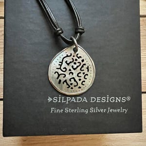 Peut inclure: Un collier avec un pendentif en argent avec un motif découpé, sur un cordon en cuir noir. Le pendentif a la forme d'une larme et présente un motif détaillé. Le collier est présenté sur une boîte noire avec le texte "SÏLPADA DESIGNS® Fine Sterling Silver Jewelry".