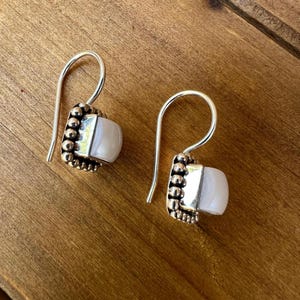 May include: A pair of silver earrings with a hook design. Each earring features a square, light-colored stone set within a beaded, bronze-colored frame. The earrings are displayed on a wooden surface.