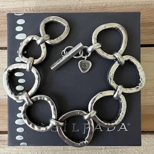 May include: A silver bracelet with large, hammered, oval-shaped links. The bracelet has a toggle clasp and a small charm with a triangle design. The bracelet is displayed on a black box with white dots.