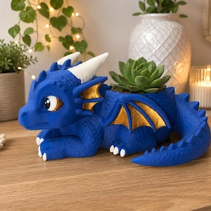 Peut inclure: Un pot de fleurs en forme de dragon bleu avec des cornes blanches et des ailes peintes en doré. La créature est en position allongée avec une plante succulente verte dans l'ouverture sur son dos. Le dragon a des écailles et de grands yeux.