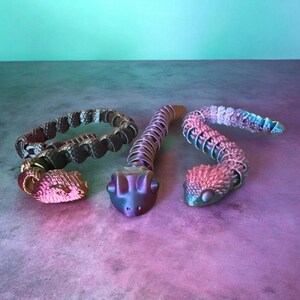 May include: Three articulated snake figurines with segmented bodies. The left snake is dark with a pink head, the centre snake is a smooth metallic purple, and the right snake features a textured, iridescent gradient of pink, blue, and purple scales.
