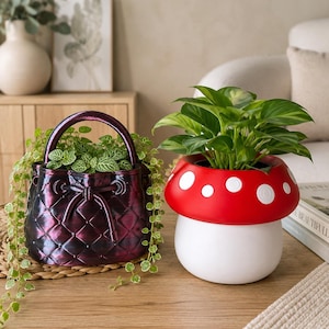 Peut inclure: Un cache-pot en forme de sac à main matelassé violet foncé avec une anse et un cordon, contenant une plante verte retombante, à côté d'un cache-pot en forme de champignon rouge et blanc abritant une plante aux larges feuilles pointues.
