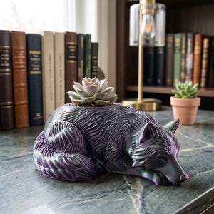 Peut inclure: Un pot de fleurs sculptural en forme de loup au repos avec une finition violette profonde et vert irisé. Le loup est allongé, la tête rentrée, et une petite plante succulente vert clair est plantée dans l'ouverture sur son dos.