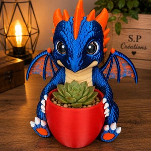 May include: A blue dragon figurine with bright orange spikes and wings, featuring large dark eyes and white claws. The dragon is holding a solid red rounded planter containing a green succulent plant with small light brown pebbles on top.