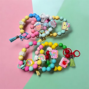 May include: Colorful beaded wrist keychains with various charms, including a mermaid, glasses, a notebook, and a rainbow. The keychains feature a mix of pastel colors, tassels, and a scissor-shaped clasp.