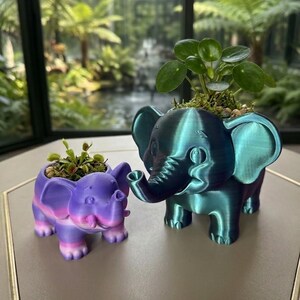 May include: Two elephant-shaped planters. The smaller planter is purple and pink with a Venus flytrap. The larger planter is a metallic teal-green colour and contains a Pilea peperomioides plant. Both planters feature four legs and trunks.