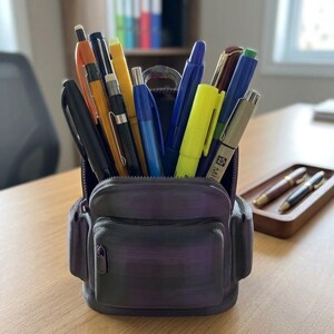 Op de afbeelding: Een donkerpaarse pennenhouder in de vorm van een miniatuurrugzak, gevuld met een assortiment pennen en stiften in diverse kleuren, waaronder zwart, oranje, geel en blauw. De houder is voorzien van een voorvak en een ritssluiting aan de bovenkant.