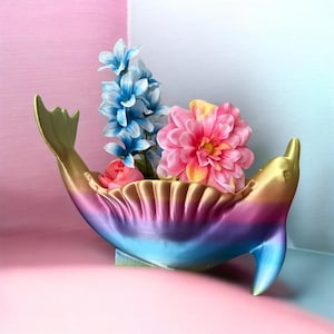 May include: A dolphin-shaped planter in gold, purple, and blue, filled with colourful artificial flowers. The planter has a tail and fin, creating a whimsical design. A decorative item for home decor, adding a touch of art.