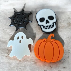 Peut inclure: Un ensemble de quatre emporte-pièces sur le thème d'Halloween comprenant une toile d'araignée noire, un crâne blanc avec des orbites et des dents noires, un fantôme blanc à l'expression surprise et une citrouille orange avec une tige.