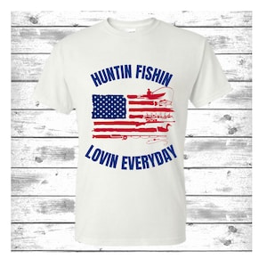 May include: White t-shirt with a blue and red American flag design. The flag has a distressed look and features hunting and fishing imagery. The text "Huntin Fishin Lovin Everyday" is printed in blue around the flag.
