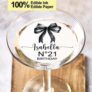 K&ouml;nnte beinhalten: Ein runder, essbarer Papiersticker in Wei&szlig; mit einer schwarzen Schleife und dem Text "Isabella N&deg;21 Birthday".