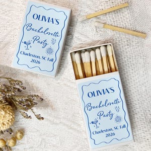 May include: Two white matchboxes with blue and white labels. The labels have the text "OLIVIA'S Bachelorette Party Charleston, SC Fall 2026".