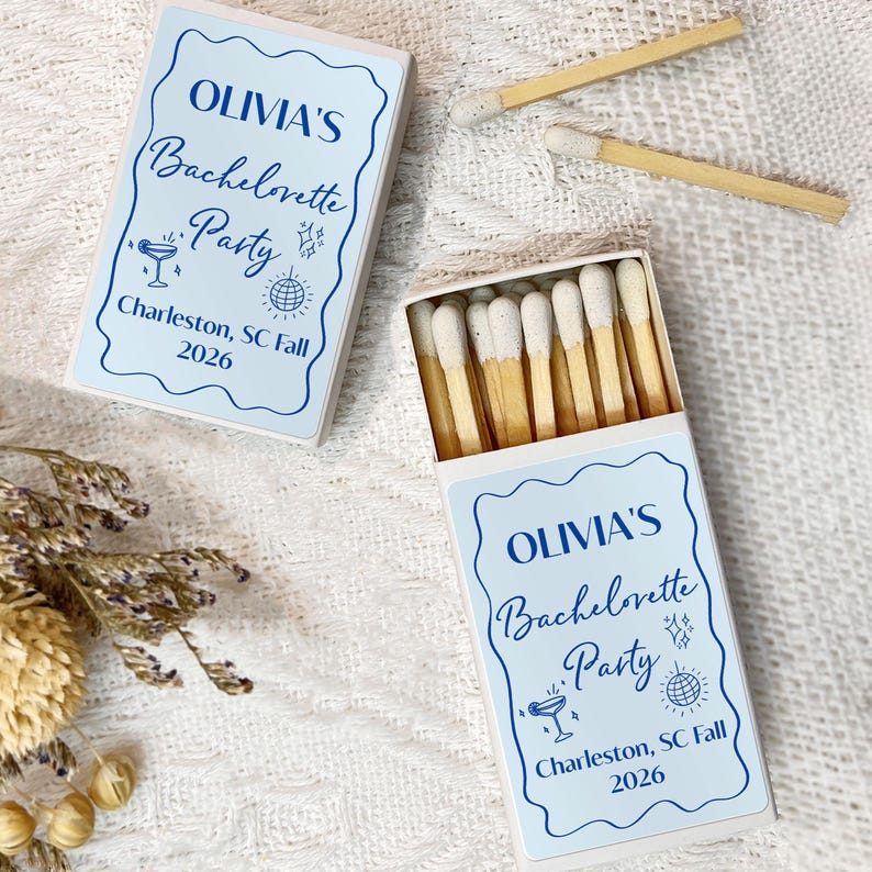 May include: Two white matchboxes with blue and white labels. The labels have the text "OLIVIA'S Bachelorette Party Charleston, SC Fall 2026".
