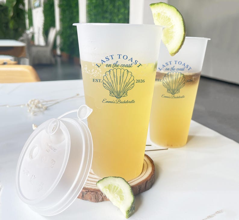 K&ouml;nnte beinhalten: Zwei durchsichtige Plastikbecher mit Deckeln, gef&uuml;llt mit einem hellgelben Getr&auml;nk. Die Becher haben ein bedrucktes Design mit dem Text "Last Toast on the Coast" und einer Muschelgrafik. Der Text "Est. 2026" und "Emma's Bachelorette" ist ebenfalls auf den Bechern aufgedruckt.