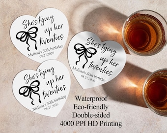May include: Heart-shaped white coasters with black text that reads "She's tying up her twenties" and "Melissa's 30th birthday 08.27.2026". Two glasses of amber liquid are visible. The coasters are waterproof, eco-friendly, and double-sided.