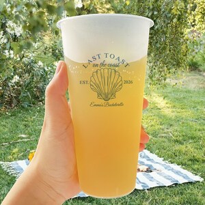 K&ouml;nnte beinhalten: Ein durchsichtiger Plastikbecher mit einem gelben Getr&auml;nk und einem wei&szlig;en Logo, auf dem "Last Toast on the Coast" mit einer Muschelgrafik und dem Text "Est. 2026" und "Emma's Bachelorette" steht.