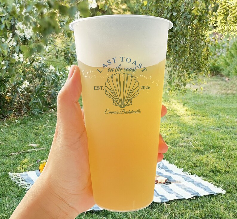 K&ouml;nnte beinhalten: Ein durchsichtiger Plastikbecher mit einem gelben Getr&auml;nk und einem wei&szlig;en Logo, auf dem "Last Toast on the Coast" mit einer Muschelgrafik und dem Text "Est. 2026" und "Emma's Bachelorette" steht.