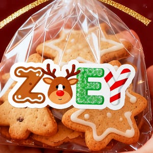 May include: A clear cellophane bag tied with a gold ribbon, filled with gingerbread cookies. The cookies are in the shapes of stars and trees, decorated with white icing. A sticker on the bag spells "ZOEY" with a gingerbread-style "Z", a reindeer, and a green "E".