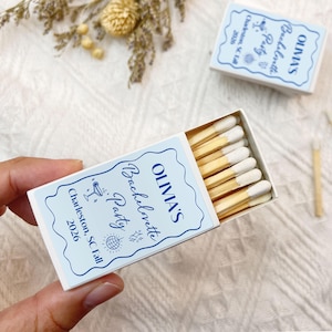 May include: White matchbox with blue and white text that reads "OLIVIA'S Bachelorette Party Charleston, SC Fall 2026". The matchbox is open and contains wooden matches with white tips.