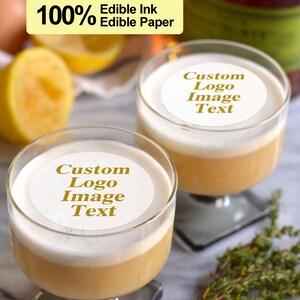 K&ouml;nnte beinhalten: Zwei klare Cocktailgl&auml;ser mit einem wei&szlig;en, runden essbaren Papierdeckel auf jedem. Die Papierdeckel haben goldenen Text, der "Custom Logo Image Text" lautet.