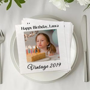 Puede incluir: Servilletas cuadradas blancas con una foto de una niña soplando las velas de un pastel de cumpleaños. El texto "Happy Birthday, Laura" y "Vintage 2019" están impresos. Las servilletas están sobre un plato blanco con cubiertos.