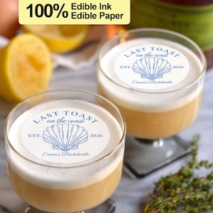 Könnte beinhalten: Zwei klare Cocktailgläser aus Glas mit einem weißen Frosting oben. Jedes Glas hat einen runden essbaren Papiersticker oben, auf dem "LAST TOAST on the coast EST. 2026 Emma's Bachelorette" mit einer blauen Muschelgrafik steht.