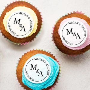 May include: Three cupcakes with white frosting and a round topper with the text "MEGAN & ANDREW" and "M&A" with the date "08.09.2025". The cupcakes are decorated with yellow, pink, and blue frosting.