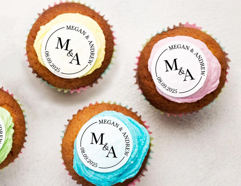 May include: Three cupcakes with white frosting and a round topper with the text "MEGAN & ANDREW" and "M&A" with the date "08.09.2025". The cupcakes are decorated with yellow, pink, and blue frosting.