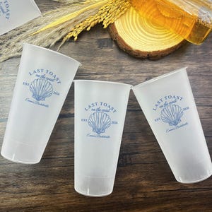 K&ouml;nnte beinhalten: Drei durchsichtige Plastikbecher mit einem blauen und wei&szlig;en Muschel-Design und dem Text "Last Toast on the Coast" mit dem Jahr 2026 und "Emma's Bachelorette" darauf gedruckt.