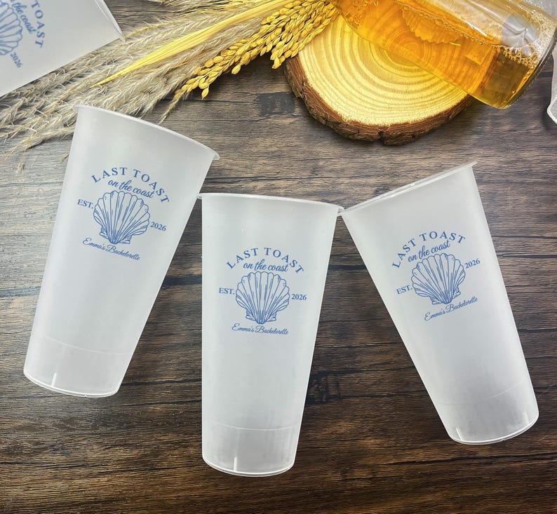 K&ouml;nnte beinhalten: Drei durchsichtige Plastikbecher mit einem blauen und wei&szlig;en Muschel-Design und dem Text "Last Toast on the Coast" mit dem Jahr 2026 und "Emma's Bachelorette" darauf gedruckt.