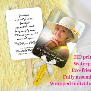 May include: A white memorial fan with a black floral design and the text "Goodbyes are not forever. Goodbyes are not the end. They simply mean I'll miss you, until we meet again."  The fan also includes the text "In loving memory of Elizabeth Thompson 17 JULY 1937 - 14 JUN 2024".  The fan is attached to a wooden stick.  The fan is next to a similar fan with a photo of an older woman wearing a yellow shirt and a white hat.  The text "In loving memory of ELIZABETH THOMPSON 17 JULY 1937 - 14 JUN 2024" is on the fan.