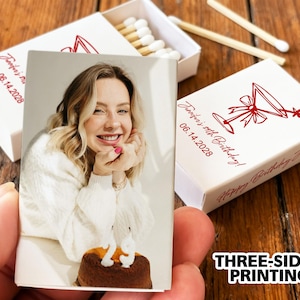 May include: A matchbox with a photo of a smiling person holding a cake. The matchbox has the text "Jennifer's 40th Birthday! 06.14.2028" and a martini glass design. The words "THREE-SIDED PRINTING" are also visible.