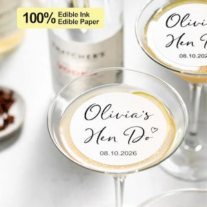 Può includere: Etichetta rotonda in carta commestibile bianca con testo nero che dice "Olivia's Hen Do" e la data "08.10.2026". L'etichetta è su un bicchiere da martini in vetro trasparente.