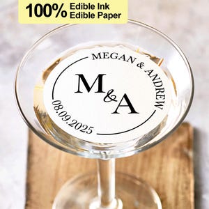May include: A white round edible paper sticker with black text that says "MEGAN & ANDREW" and "M&A" with a date of "08.09.2025" in a circle. The sticker is on a clear glass martini glass.