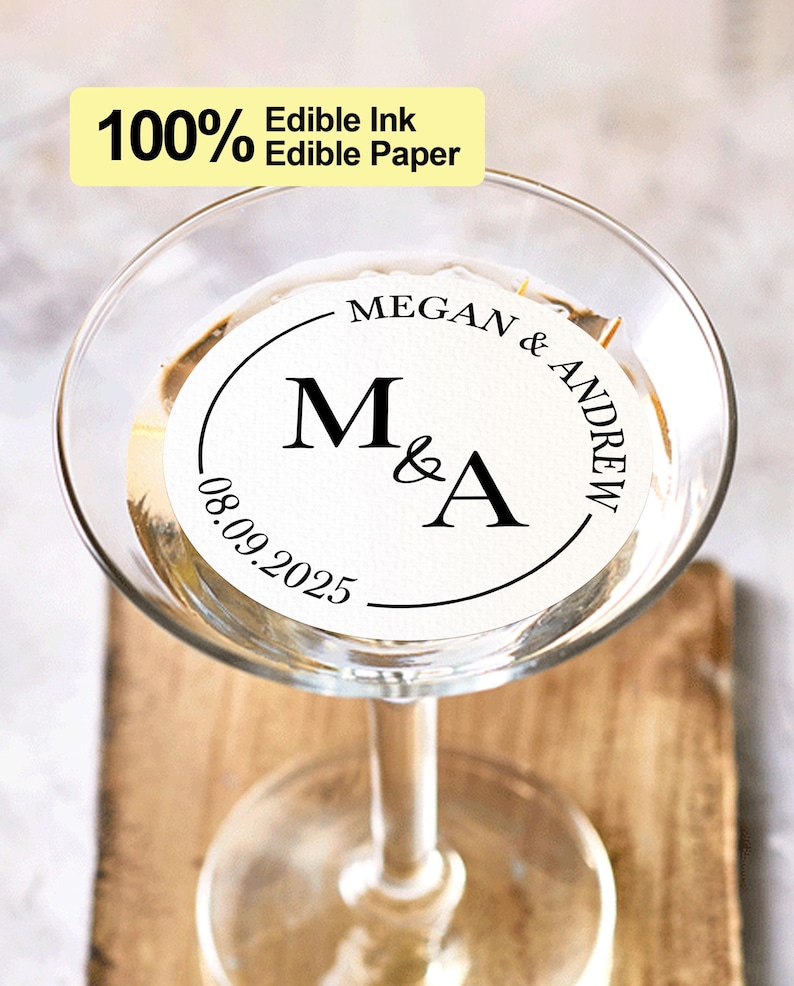 May include: A white round edible paper sticker with black text that says "MEGAN & ANDREW" and "M&A" with a date of "08.09.2025" in a circle. The sticker is on a clear glass martini glass.
