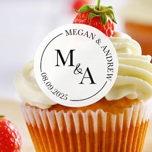 May include: White cupcake with white frosting and a round sticker that says "Megan & Andrew" with the initials "M&A" and the date "08.09.2025".