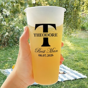 Può includere: Un bicchiere di plastica trasparente con una bevanda gialla al suo interno. Il bicchiere ha un design nero con la lettera "T" e il testo "Theodore Best Man 08.07.2026".