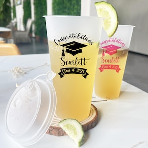 May include: Two clear plastic cups with lids and straws. The cups are decorated with black and pink graduation caps and the text "Congratulations Scarlett Class of 2023".