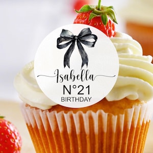 K&ouml;nnte beinhalten: Ein wei&szlig;er Cupcake mit wei&szlig;em Frosting und einem runden Aufkleber in Schwarzwei&szlig;, auf dem "Isabella N&deg;21 Birthday" mit einer schwarzen Schleife steht.