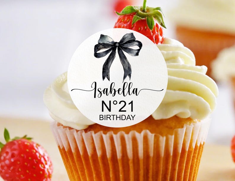 K&ouml;nnte beinhalten: Ein wei&szlig;er Cupcake mit wei&szlig;em Frosting und einem runden Aufkleber in Schwarzwei&szlig;, auf dem "Isabella N&deg;21 Birthday" mit einer schwarzen Schleife steht.