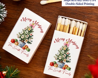 May include: Two white matchboxes with a Christmas tree design. The tree is decorated with Mahjong tiles and topped with a pink bow. The text "Merry Mahjong" and "The Smith Family" are printed on the boxes. The matches are visible in one box.