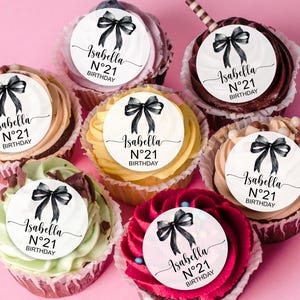 K&ouml;nnte beinhalten: Sechs Cupcakes, die mit runden essbaren Bildern verziert sind, die einen schwarzen Bogen und den Text "Isabella N&deg;21 BIRTHDAY" zeigen.